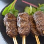 Kofta on wooden skewers with green leafy garnish and a bowl of tahini sauce arranged on a black plate.