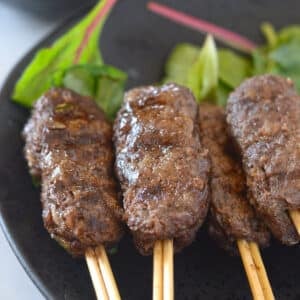 Kofta on wooden skewers with green leafy garnish and a bowl of tahini sauce arranged on a black plate.