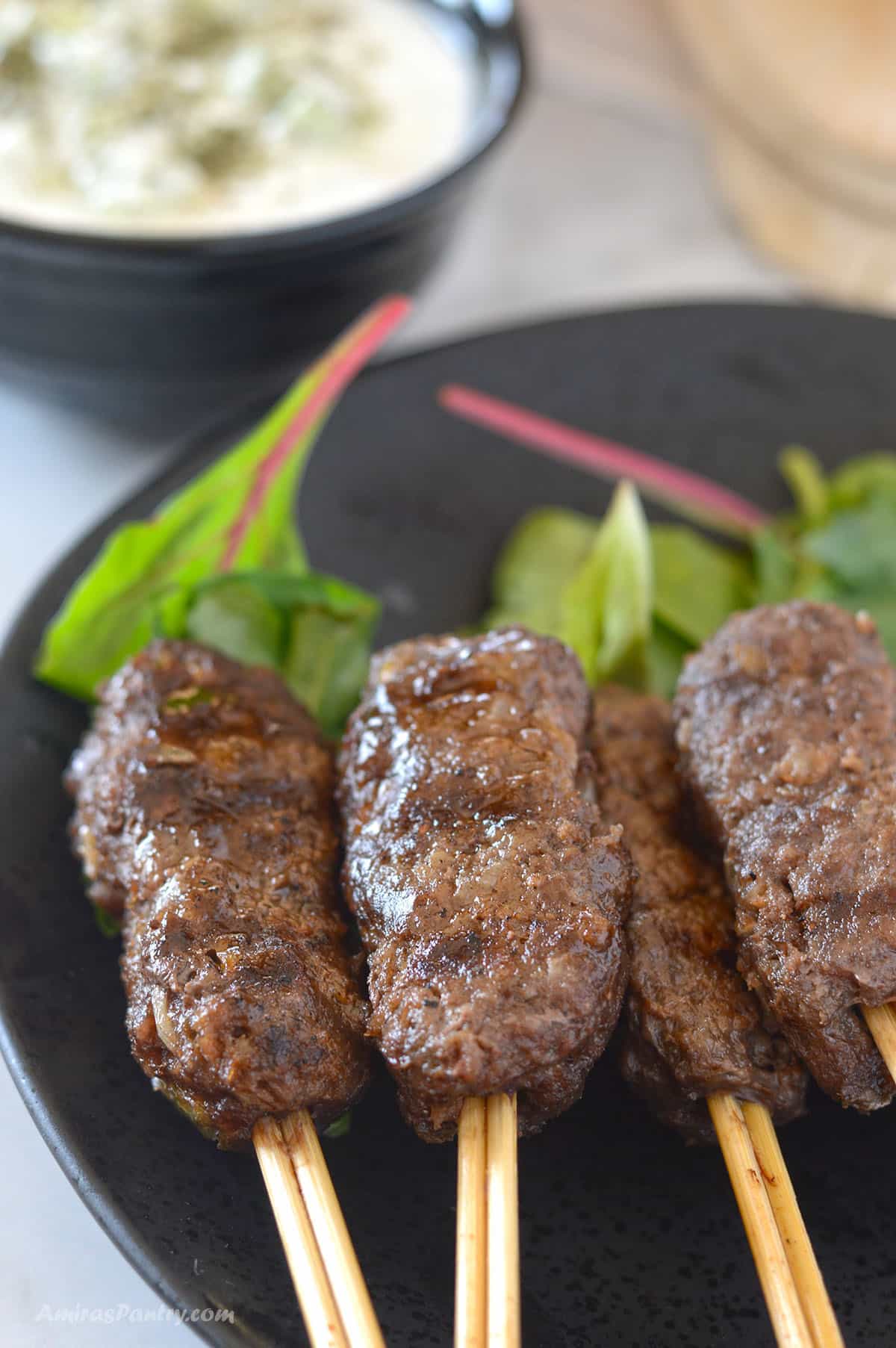 Four Kofta kebabs on wooden skewers with green leafy garnish arranged on a black plate. 