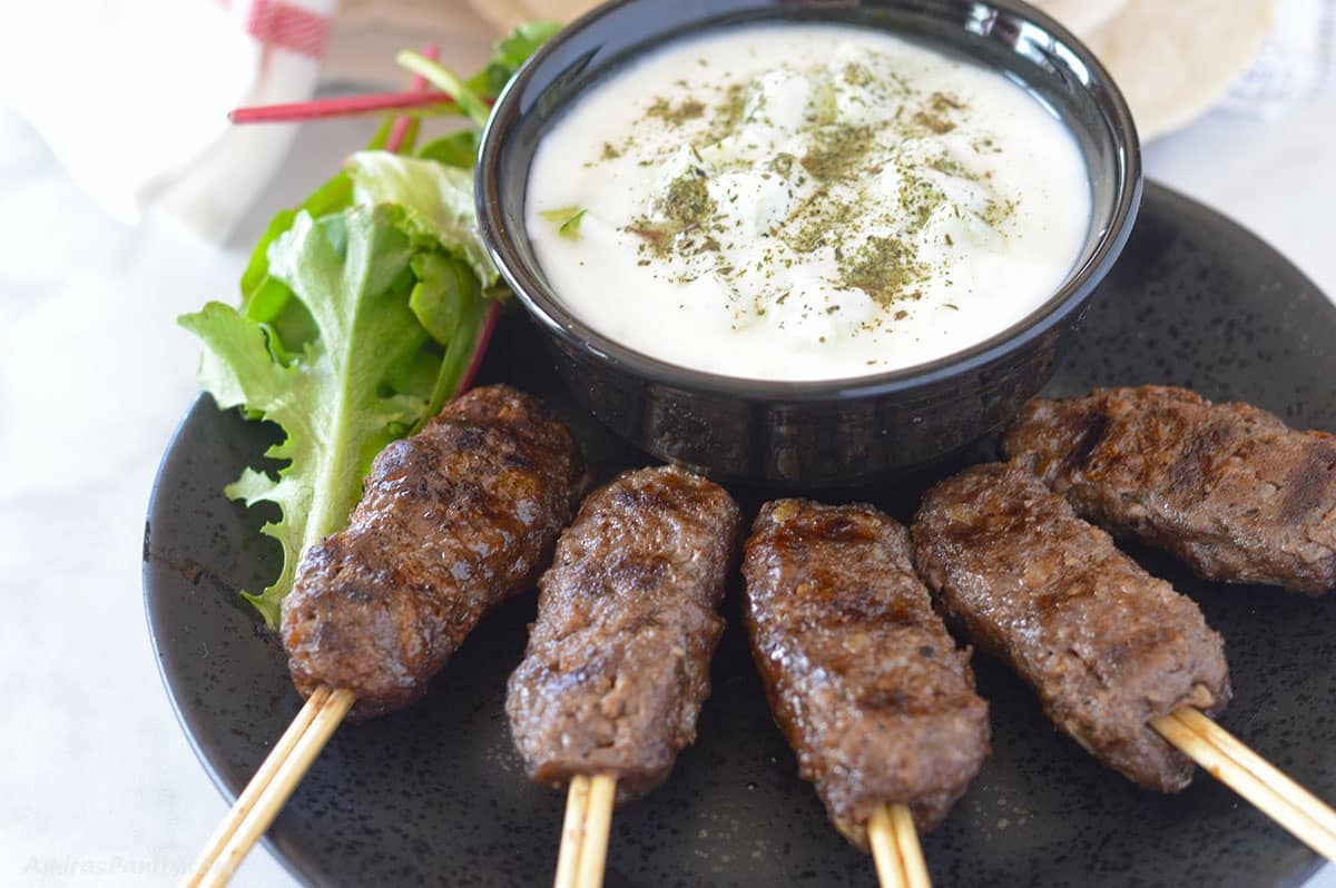 Grilled kofta kebabs on wooden skewers are arranged in a semi-circle on a black plate. Positioned above them on the same plate is a black bowl filled with a creamy yogurt cucumber sauce sprinkled with dried herbs.