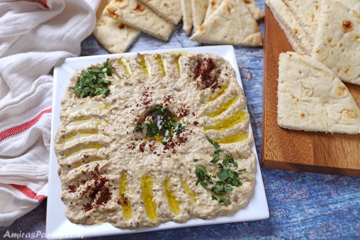 Overhead view of mutabal eggplant dip on a square white plate, drizzled with olive oil and garnished with chopped parsley and sumac, surrounded by triangle-cut pita bread on a wooden board.