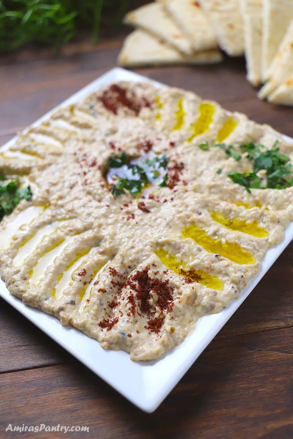 Mutabal eggplant dip spread on a square white plate, drizzled with olive oil and topped with chopped parsley and sumac, served with triangle-cut pita bread.