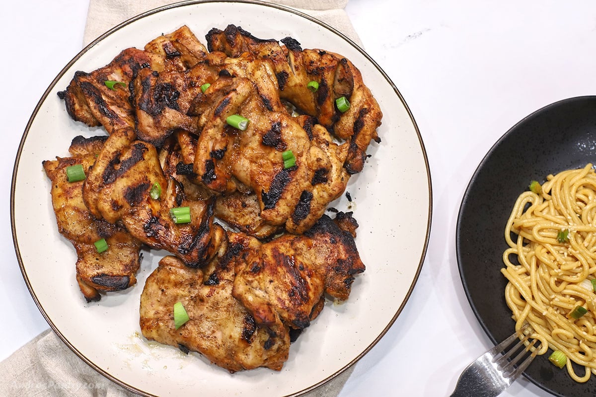Overhead view of grilled soy sauce chicken thighs piled on a white plate, garnished with chopped green onions, next to a black bowl of sesame noodles.