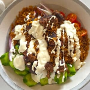 Close up overhead view of a crispy rice salad bowl with spiced ground meat, creamy white sauce, cucumbers, cherry tomatoes, and red onion.
