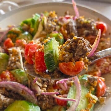 A close up shot of a spoon lifting a bite of Crispy Rice Salad from a bowl, showcasing ground beef chunks, cucumbers, tomatoes, red onions, and golden crispy rice coated in a creamy white sauce.
