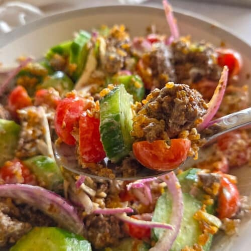 A close up shot of a spoon lifting a bite of Crispy Rice Salad from a bowl, showcasing ground beef chunks, cucumbers, tomatoes, red onions, and golden crispy rice coated in a creamy white sauce.