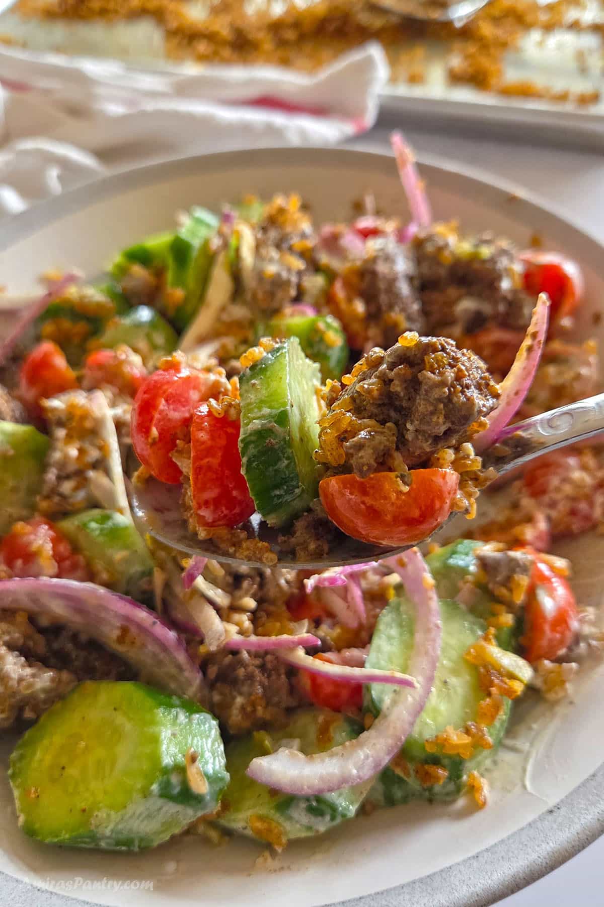 A close up shot of a spoon lifting a bite of Crispy Rice Salad from a bowl, showcasing ground beef chunks, cucumbers, tomatoes, red onions, and golden crispy rice coated in a creamy white sauce.