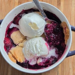 Close-up overhead shot of a two-ingredient berry cobbler in a small white dish with handles, topped with two scoops of melting vanilla ice cream and a spoon resting inside