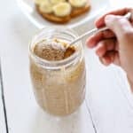 A hand scooping a spoonful of creamy almond butter from a mason jar, with banana toast visible on a plate in the background.