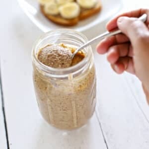 A hand scooping a spoonful of creamy almond butter from a mason jar, with banana toast visible on a plate in the background.