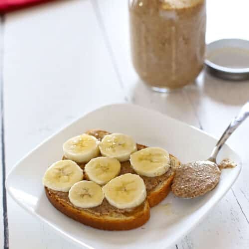Toast spread with almond butter and topped with banana slices, served on a white plate with a spoonful of almond butter alongside and a mason jar of almond butter in the background.