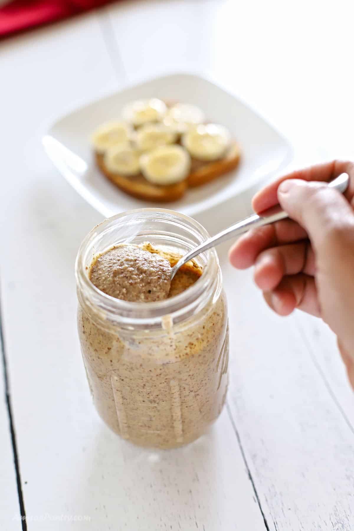 A hand scooping almond butter from a mason jar, with a slice of toast topped with banana rounds visible on a white plate in the background.