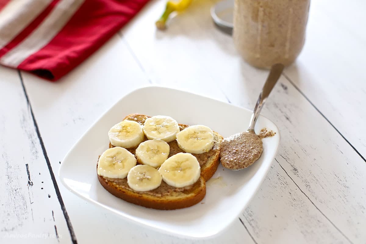 Almond butter toast topped with banana slices on a white plate, with a spoonful of almond butter resting on the plate and a mason jar of almond butter in the background.