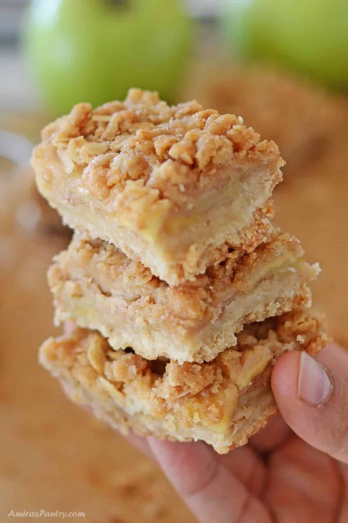 A hand holding a stack of buttery apple crisp bars with cinnamon apples.