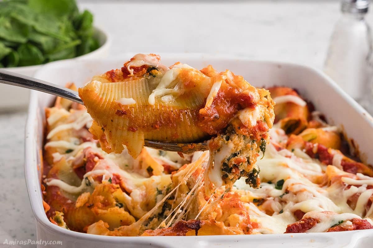 A serving spoon lifting a cheesy, baked stuffed conchiglioni shell from a pan, showing stretchy melted mozzarella cheese.