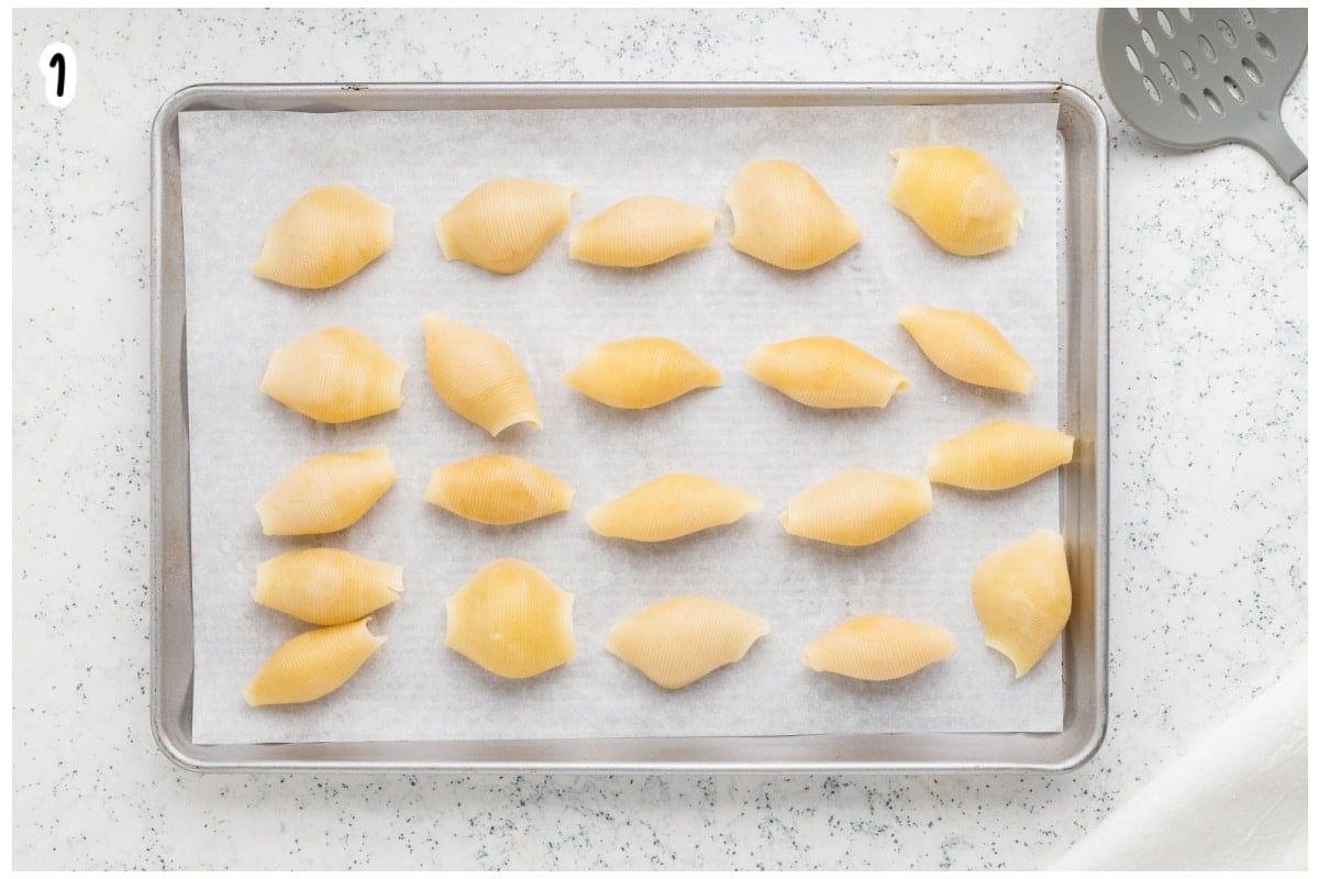 Cooked jumbo pasta shells laid out in rows on a parchment lined baking sheet to cool, labeled as step 1.