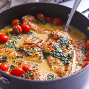 Close-up of finished creamy Italian chicken with wilted spinach and cherry tomatoes in a golden cream sauce, served in a skillet with a spoon.