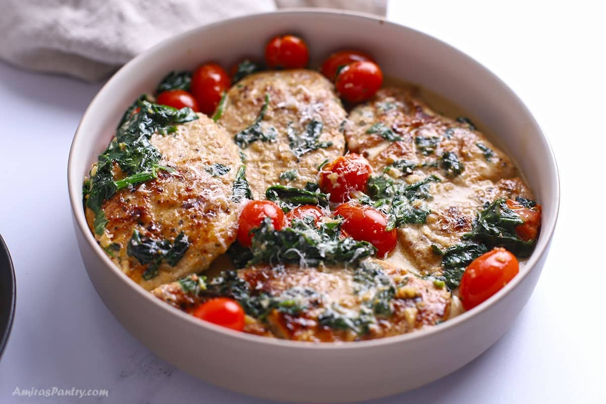 Creamy Italian chicken breasts topped with spinach, cherry tomatoes, and grated Parmesan, served in a beige bowl.