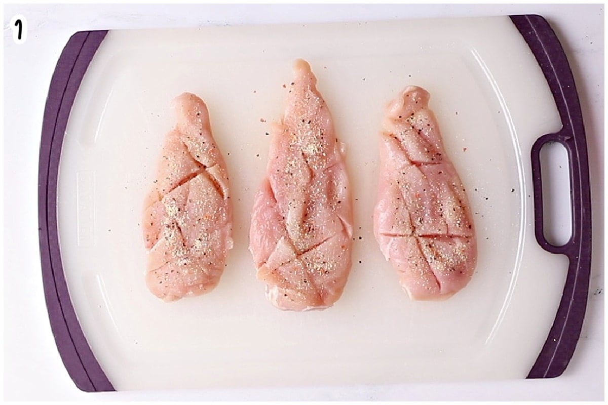 Three raw chicken breasts scored with shallow crosshatch cuts and seasoned with Italian dressing seasoning on a cutting board.