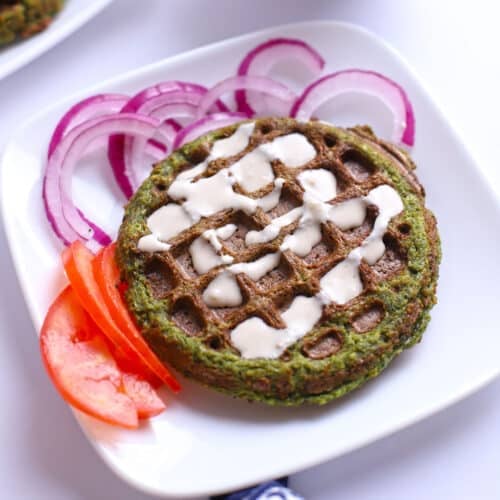 Close-up overhead view of a round falafel waffle drizzled with tahini sauce, served on a white plate with sliced red onions and fresh tomato wedges.