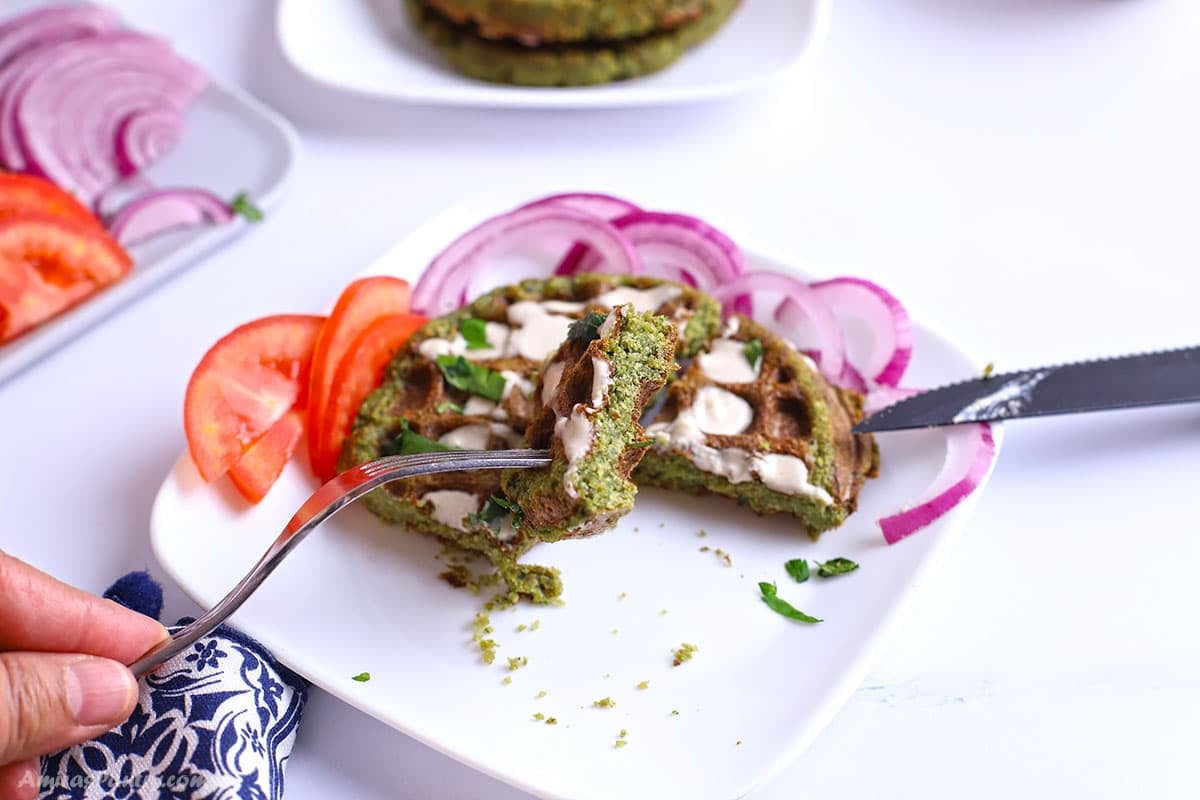 A hand using a fork and knife to cut into a tahini-drizzled falafel waffle garnished with chopped parsley.