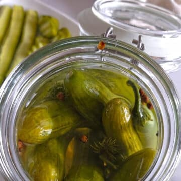 Close-up overhead view of an open glass jar filled with quick pickled cucumbers, dill, and jalapeรฑo in brine, with cucumber spears on a plate in the background.