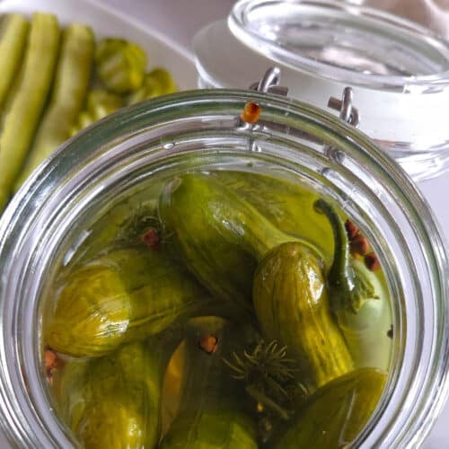 Close-up overhead view of an open glass jar filled with quick pickled cucumbers, dill, and jalapeño in brine, with cucumber spears on a plate in the background.