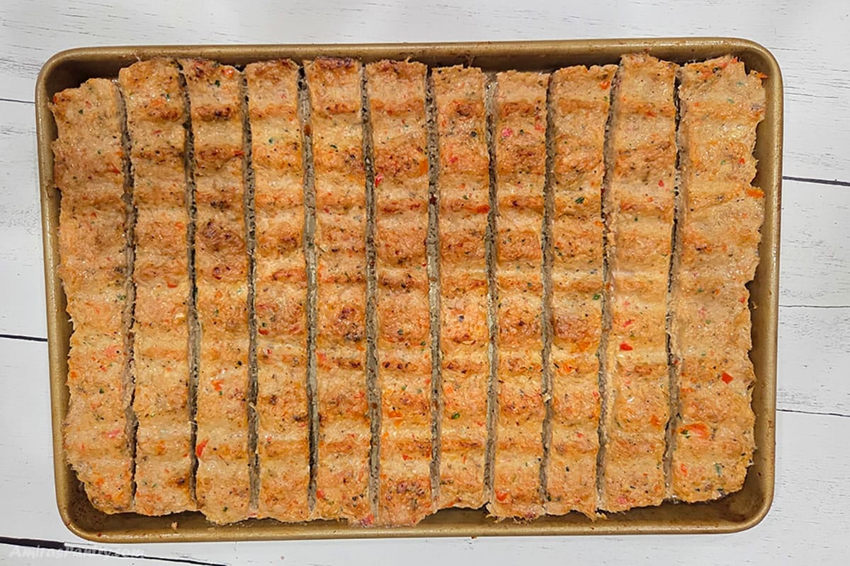 Overhead view of baked sheet pan kebab strips scored into rows on a gold baking pan, with a golden-brown crust and visible red pepper and herb flecks.