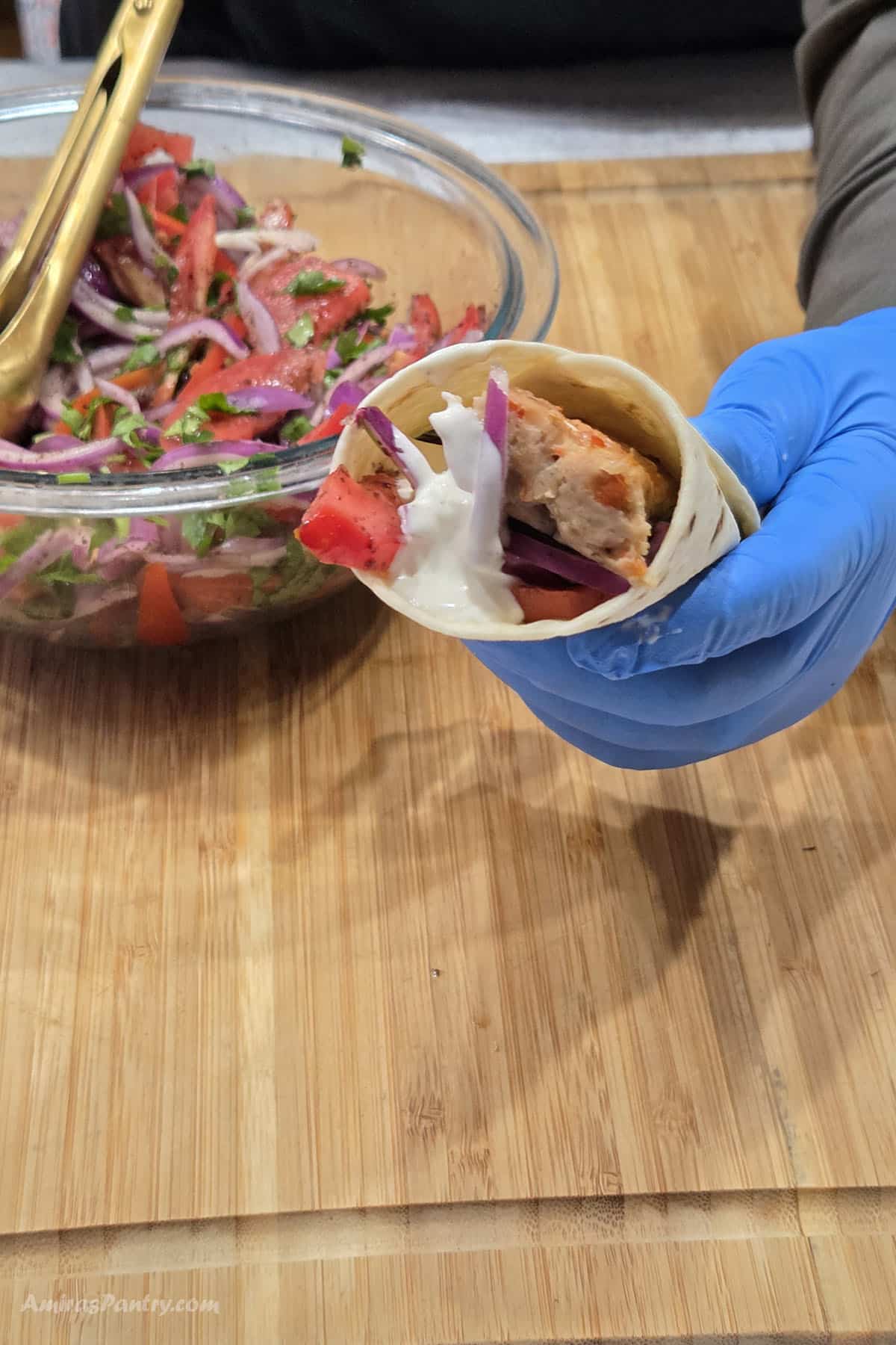 A hand holding a flatbread wrap filled with chicken kebab, tomato, red onion, and white sauce, with a bowl of fresh vegetable salad in the background.