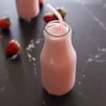 A single glass milk bottle filled with strawberry banana smoothie and a red straw, with fresh strawberries and a second smoothie bottle visible in the background.