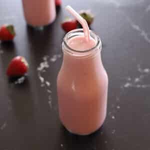 A single glass milk bottle filled with strawberry banana smoothie and a red straw, with fresh strawberries and a second smoothie bottle visible in the background.