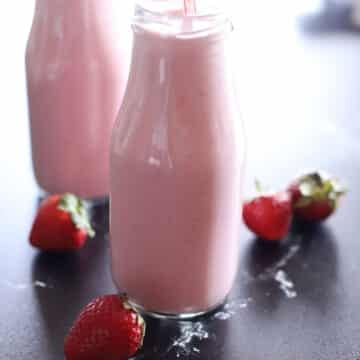 Close-up of two glass milk bottles filled with a creamy pink strawberry-banana smoothie, with fresh whole strawberries scattered around the base on a dark surface.
