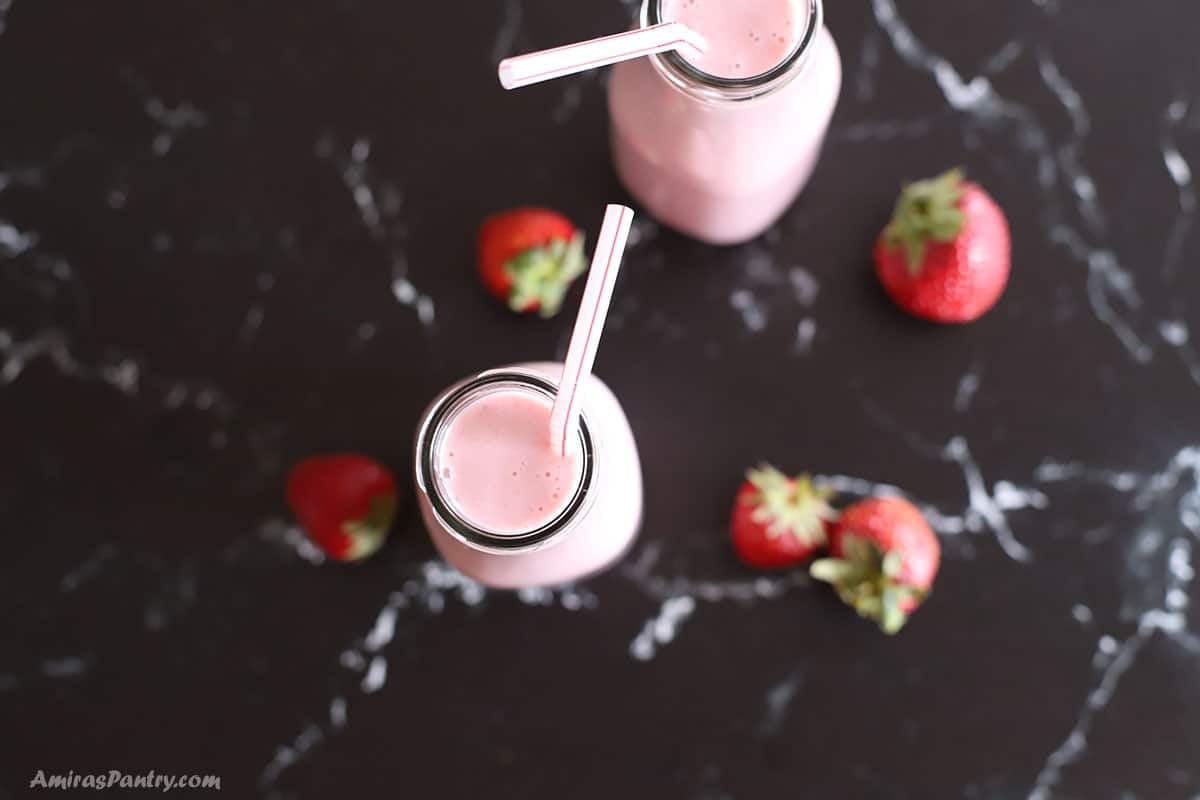 Overhead view of two glass milk bottles filled with creamy pink strawberry-banana smoothie and striped pink straws, surrounded by fresh whole strawberries on a dark marble surface.