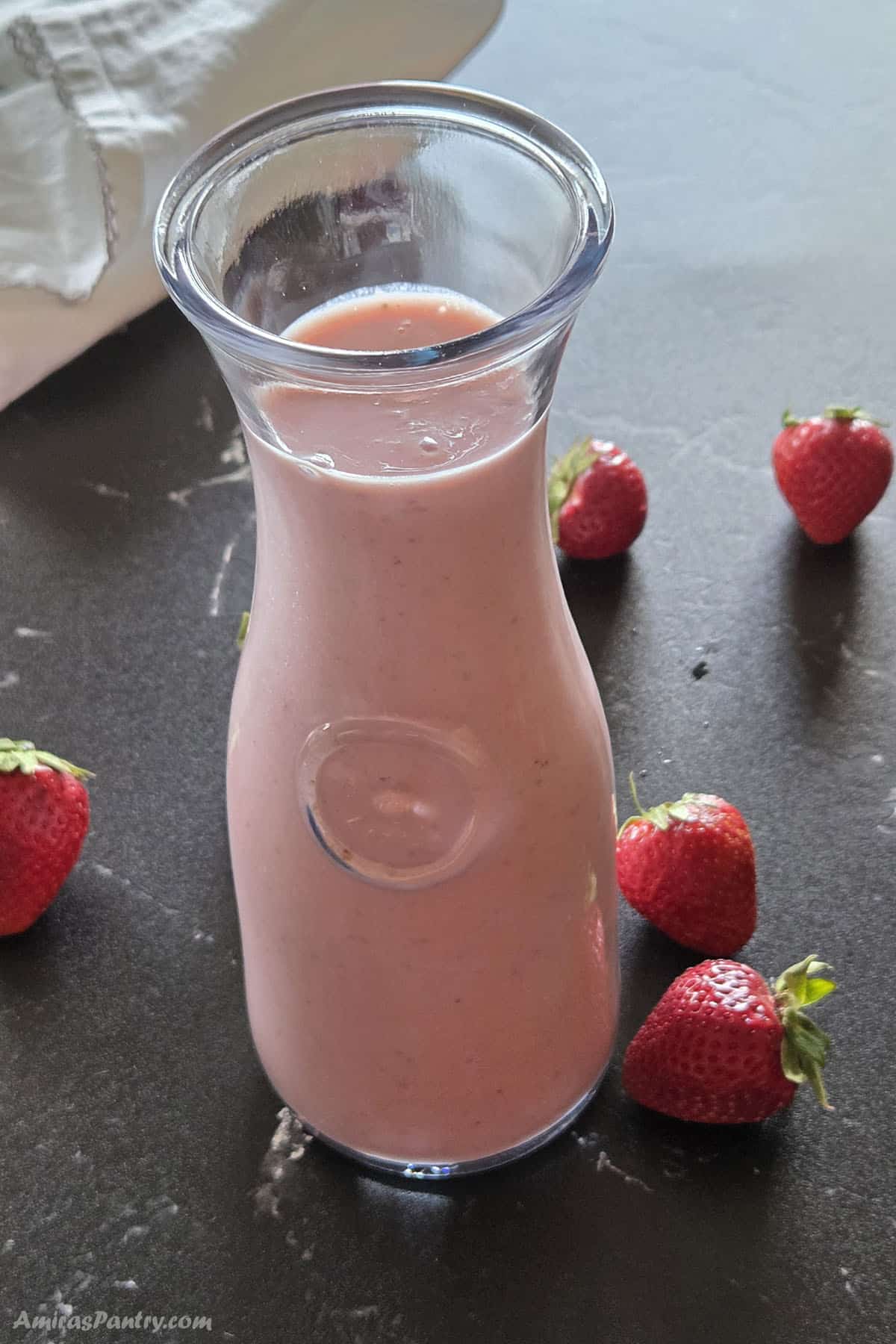 A clear glass carafe filled with a creamy, pink strawberry banana smoothie, surrounded by five scattered whole fresh strawberries on a dark countertop.