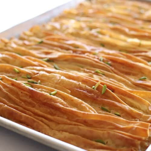 Close-up angled view of golden-brown crinkled phyllo pastry strips in a baking tray, glossy with butter and sprinkled with chopped pistachios.