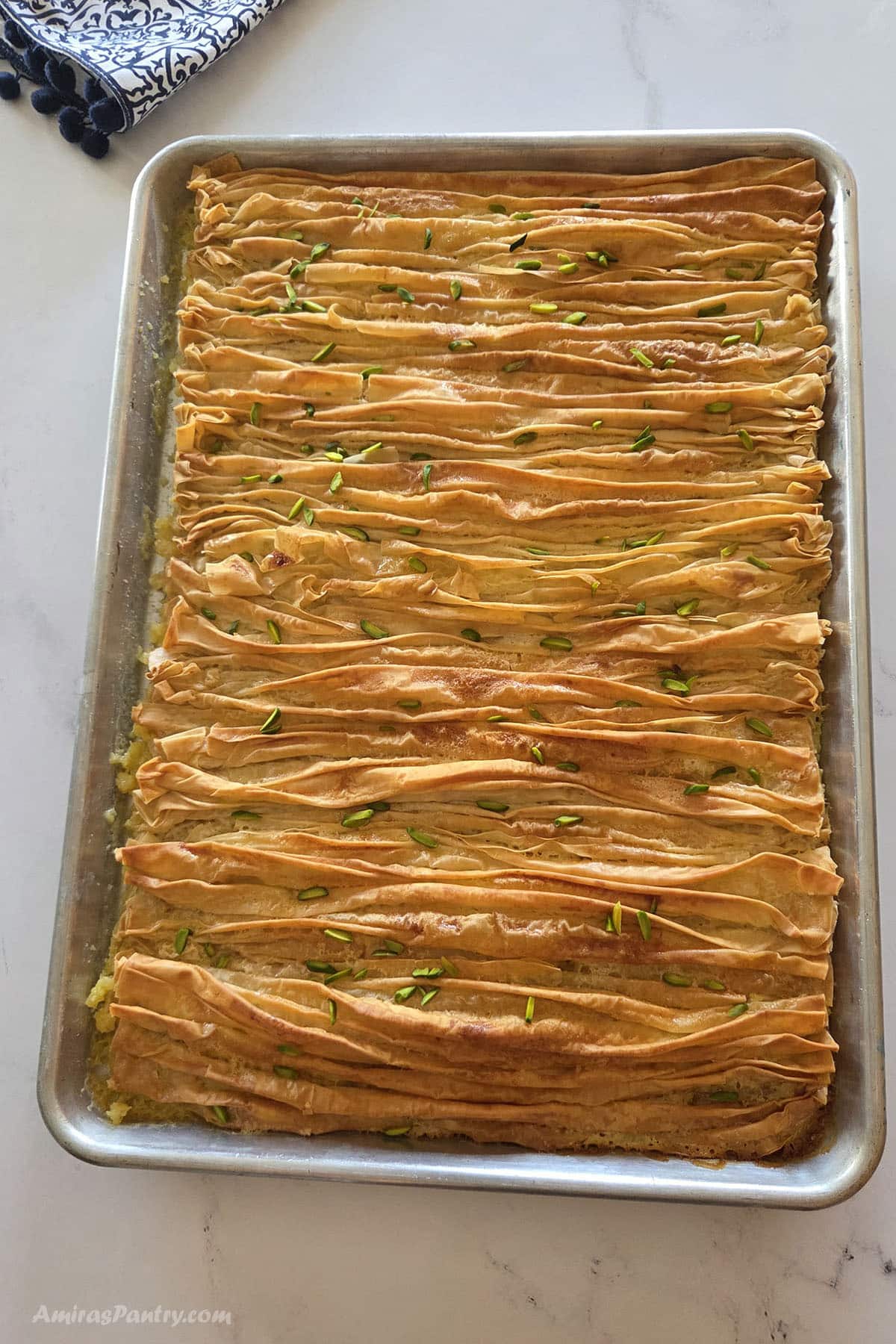 A baking tray filled with crinkled phyllo pastry strips arranged in rows, baked golden brown and topped with chopped pistachios, on a marble countertop.