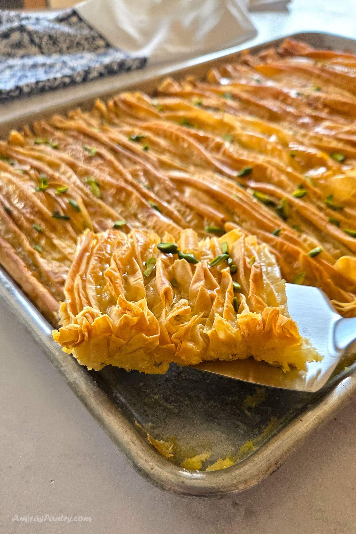 A metal spatula lifting a slice of golden crinkled phyllo pastry out of the baking tray, showing its ruffled layers and pistachio garnish, with more of the pastry in the background.