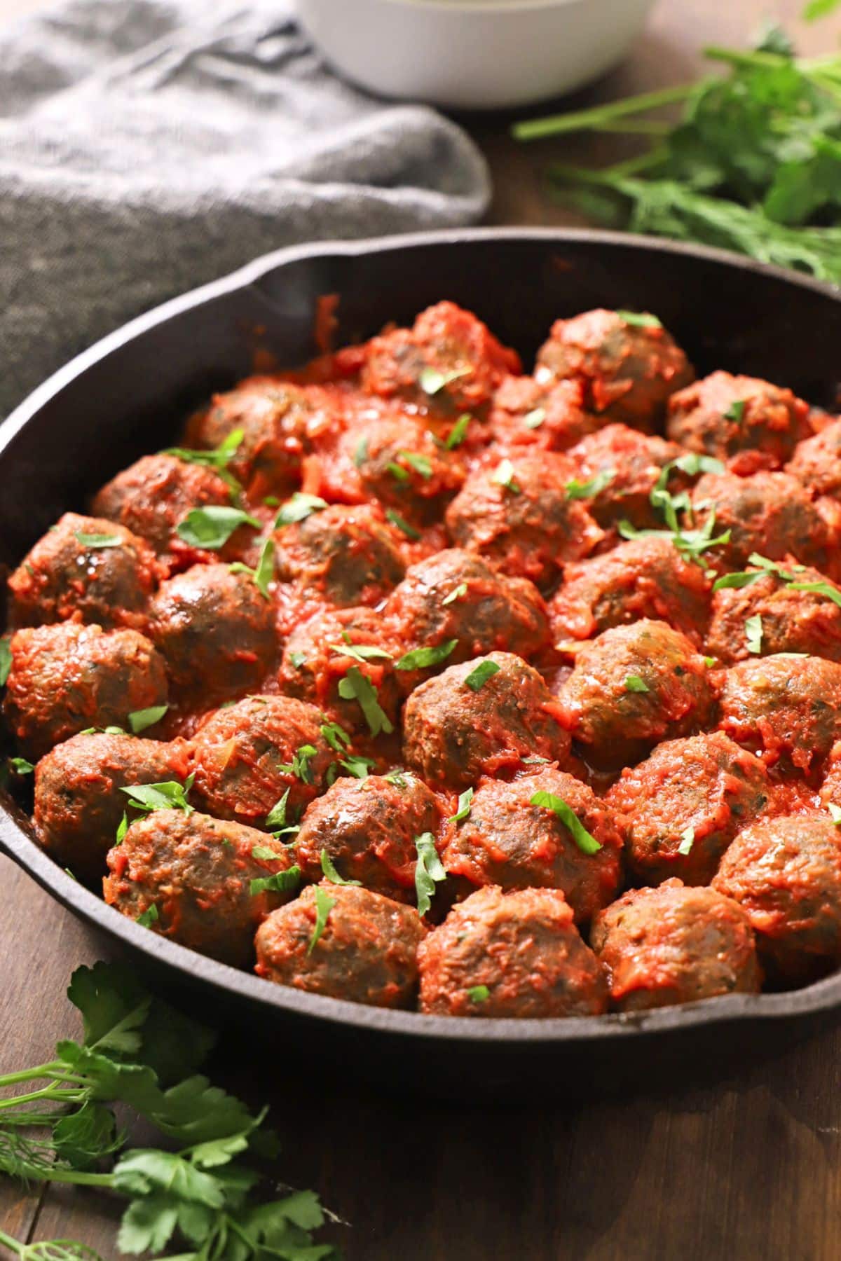 A dark skillet filled with rice meatballs simmered in tomato sauce and garnished with chopped parsley, set on a wooden table with fresh herbs and a bowl in the background.