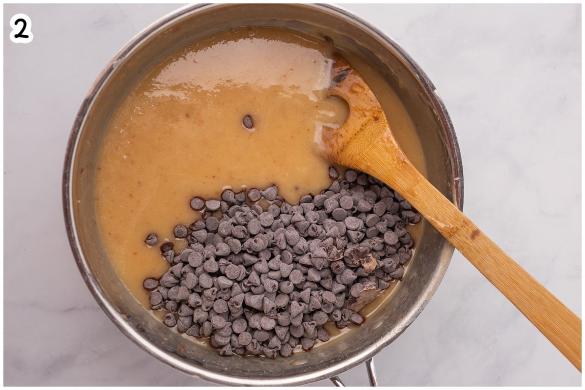 Overhead view of a saucepan with chocolate chips being added to hot melted butter, sugar, and evaporated milk mixture, with a wooden spoon resting inside.