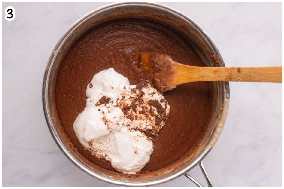 Overhead view of marshmallow fluff being added to melted chocolate fudge mixture in a saucepan, with a wooden spoon resting inside.