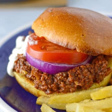 Close-up of a ground beef shawarma sandwich topped with tomato, red onion, and a creamy sauce on a bun, served with fries on a blue plate.