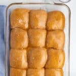 Close up overhead shot of golden whole wheat dinner rolls in a glass baking dish, glossy with butter and topped with flaky salt, with a blue linen napkin visible along the edge of the frame.