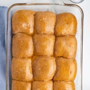 Close up overhead shot of golden whole wheat dinner rolls in a glass baking dish, glossy with butter and topped with flaky salt, with a blue linen napkin visible along the edge of the frame.