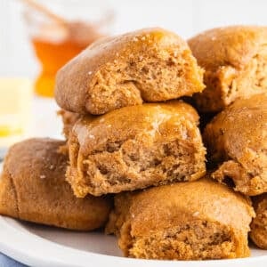 Close-up of stacked whole wheat dinner rolls on a white plate, some torn open to show the soft, fluffy interior, with a jar of honey and butter blurred in the background.
