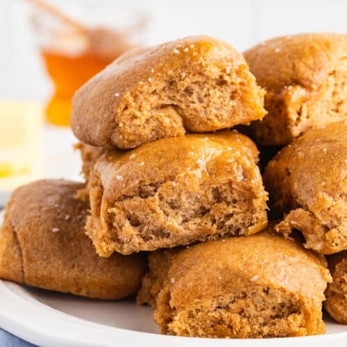 Close-up of stacked whole wheat dinner rolls on a white plate, some torn open to show the soft, fluffy interior, with a jar of honey and butter blurred in the background.