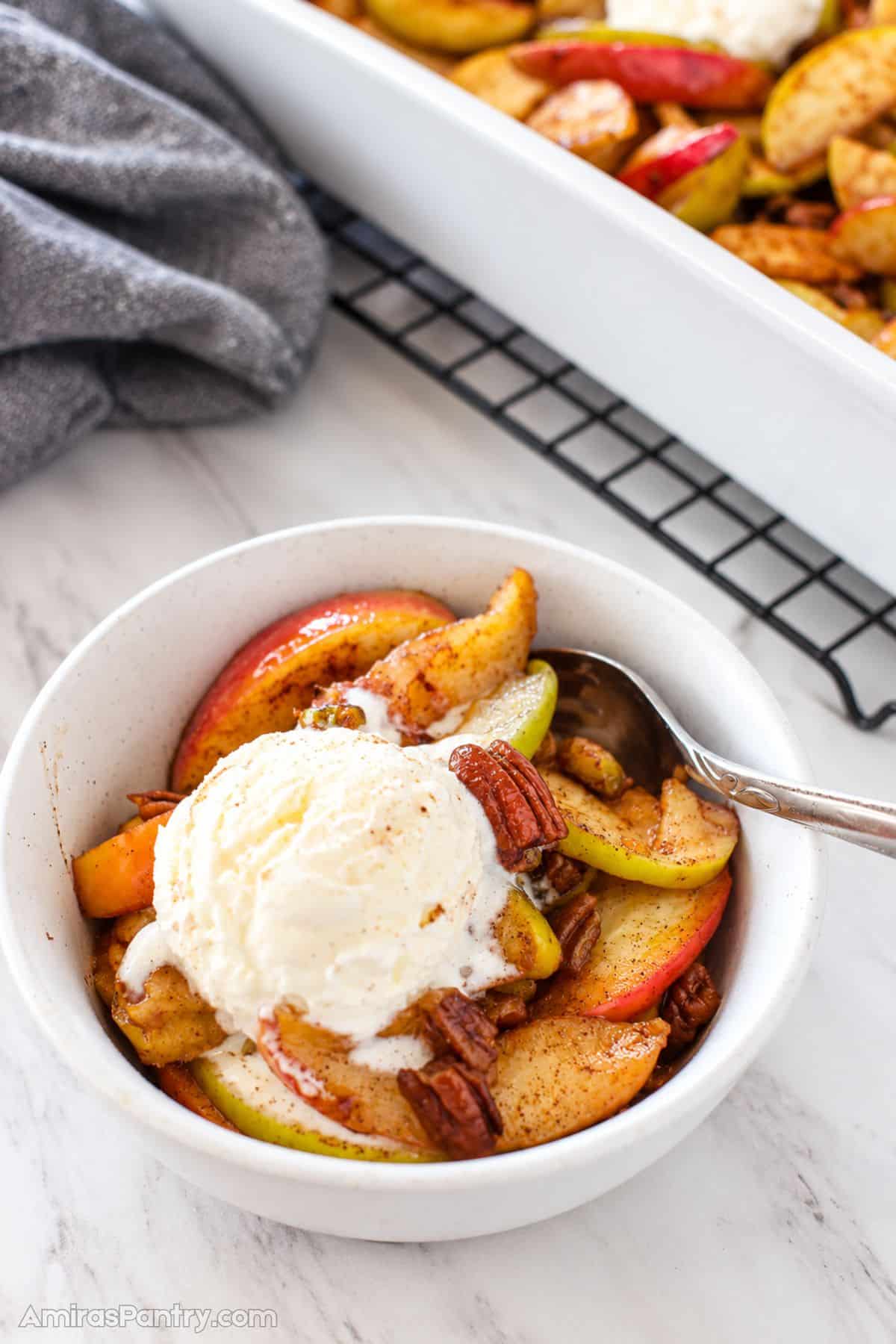 Bowl of warm baked apple slices with pecans, topped with a melting scoop of vanilla ice cream, with the baking dish of remaining apples in the background.