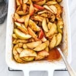 Close up of cinnamon spiced baked apple slices with pecans in a white baking dish, with a spoon lifting sauce from the corner.