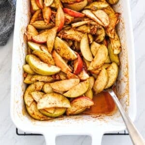 Close up of cinnamon spiced baked apple slices with pecans in a white baking dish, with a spoon lifting sauce from the corner.