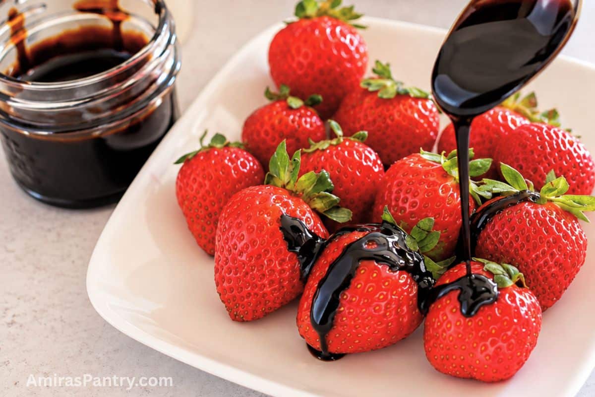Balsamic glaze being drizzled from a spoon over fresh strawberries on a white plate, with a jar of glaze beside it.