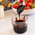 A silver spoon drizzling thick, glossy balsamic glaze into a small glass jar, with a bowl of fresh strawberries in the background.